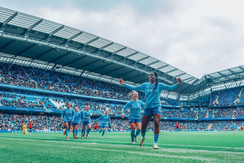 El City pone la liga al rojo y pinta Manchester de azul. Lauren Hemp y varias jugadoras del City corren a felicitar a Bunny Shaw tras la victoria. Al fondo, la grada del Etihad abarrotada de aficionados.