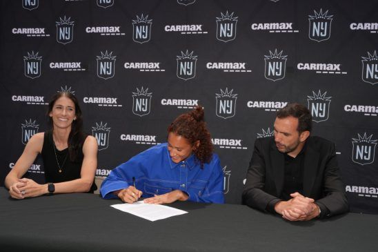 Lynn Williams seguirá en Gotham. A la izquierda, Yael Averbuch West, directora general del club. A la derecha, Juan Carlos Amorós. entrenador del equipo. Ambos miran atentos y sonrientes cómo la jugadora, en el centro, firma su nuevo contrato.
