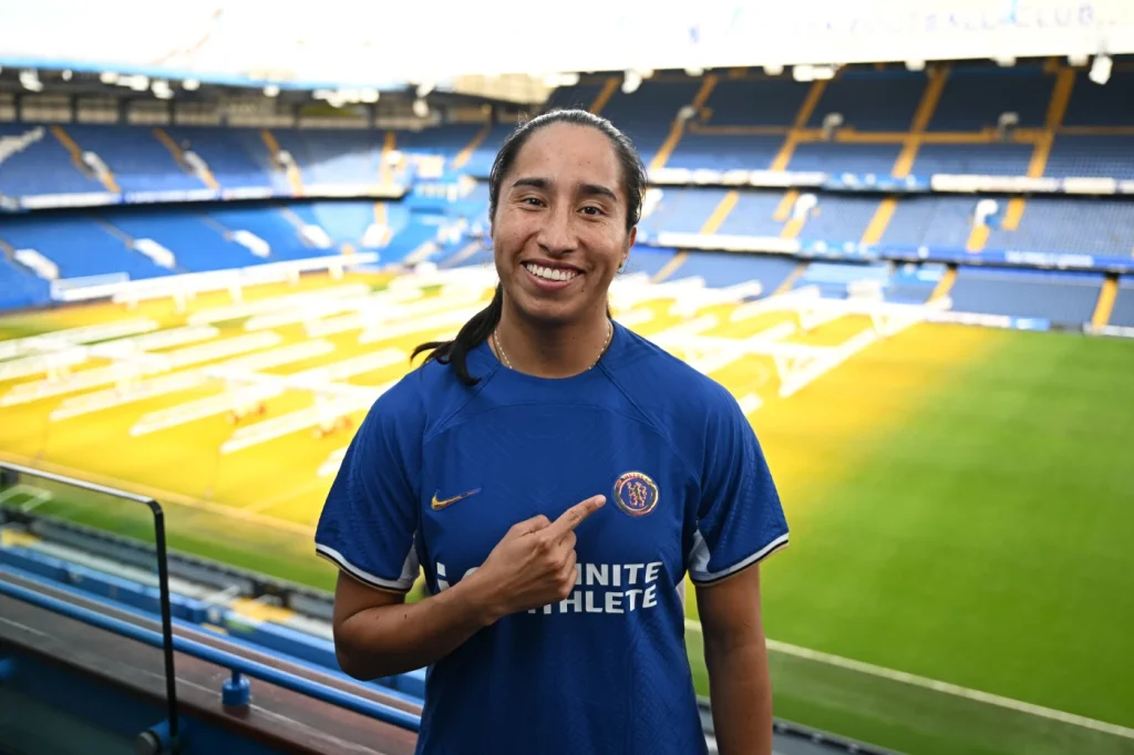 Mayra Ramírez hace historia fichando por el Chelsea. La jugadora posa señalándose el escudo de su nueva camiseta desde la gradería de Stamford Bridge.