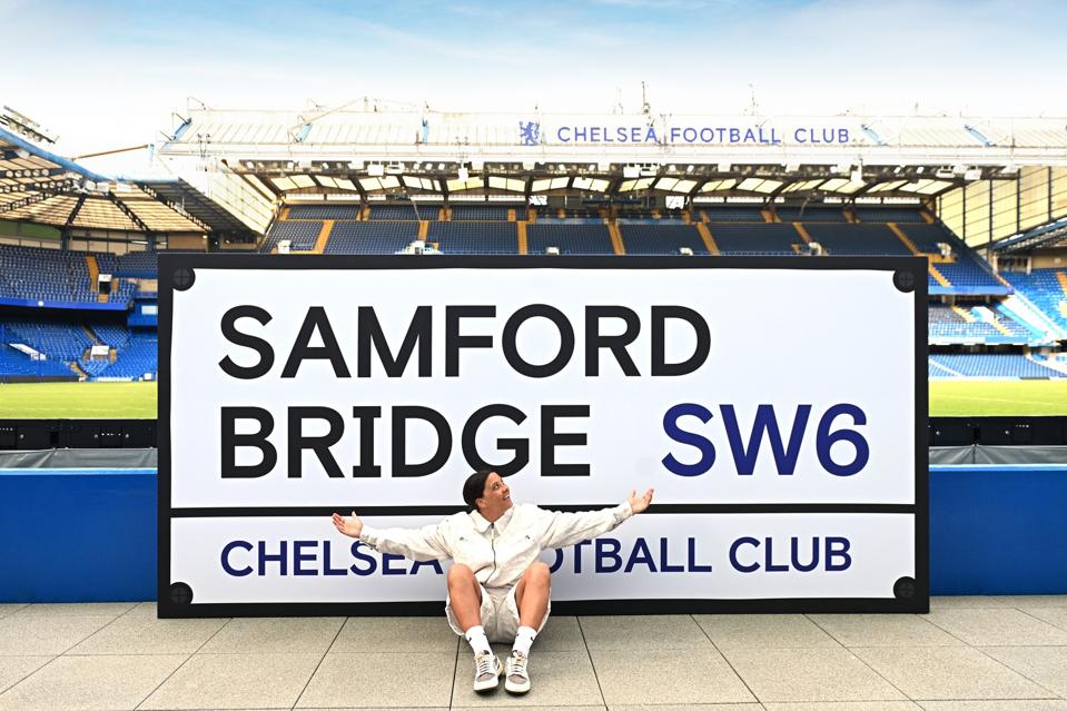 Sam se Kerrda. Sam Kerr posa en Stamford Bridge con una señal callejera tamaño XXL que reza: "SAMFORD BRIDGE".